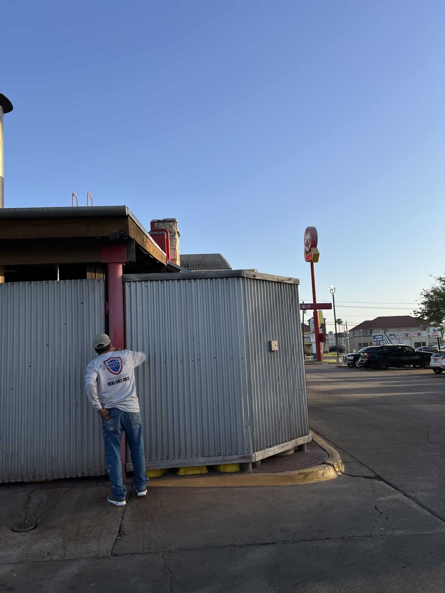 A man in a cap and jeans stands by a corrugated metal building under a clear blue sky. In the distance is a fast-food sign and parked cars, evoking a calm morning scene.