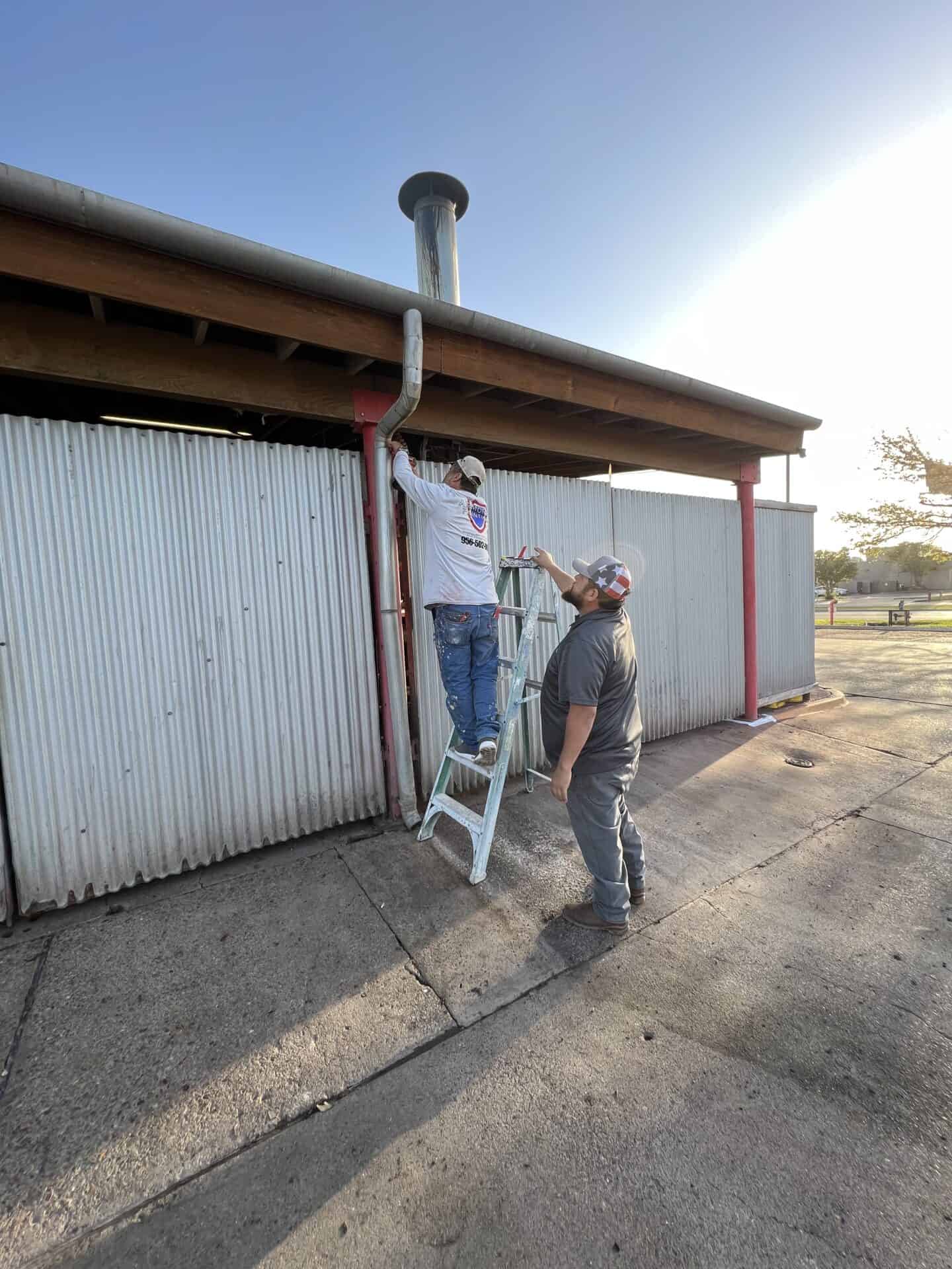 Two men work on a corrugated metal building. One stands on a ladder adjusting a pipe, while the other holds the ladder. The setting sun casts long shadows.