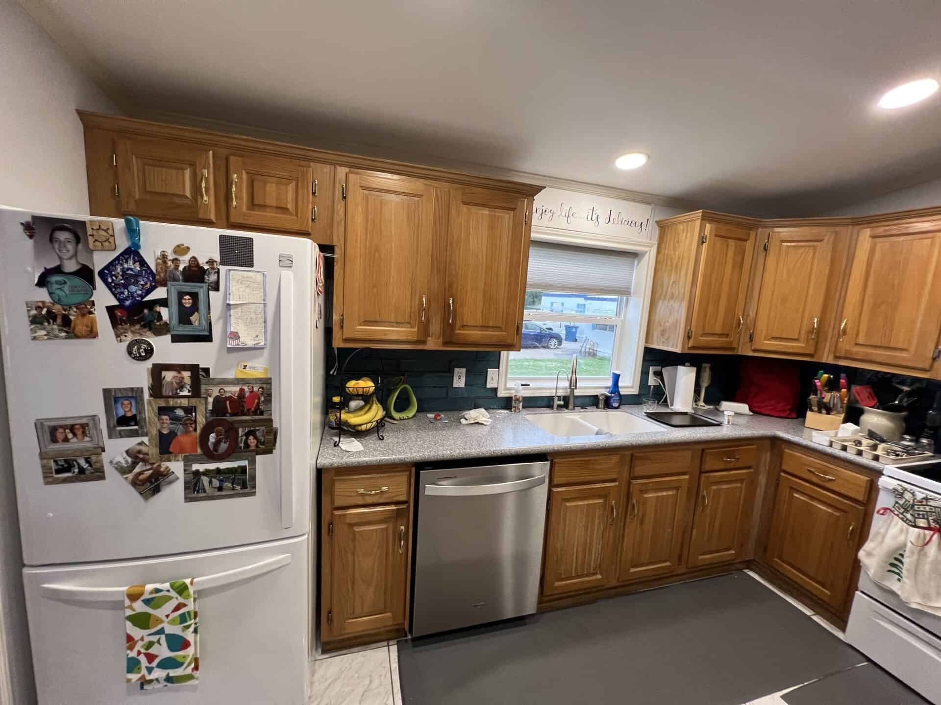 Cozy kitchen with wooden cabinets, a fridge adorned with family photos, and a window above the sink. Warm, inviting atmosphere with personal touches.