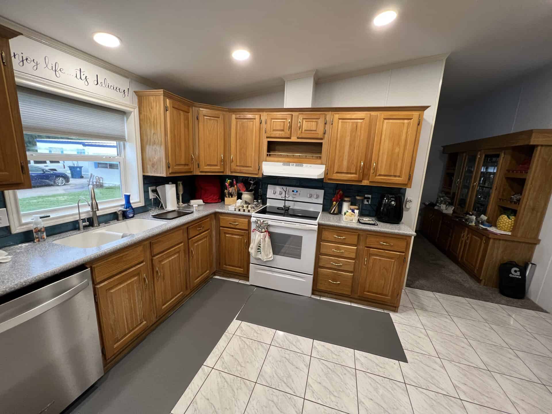 Warm, inviting kitchen with wooden cabinets and stainless steel appliances, including a stove and dishwasher. A large window overlooks the outdoors, and a sign above reads, "Enjoy life... it's delicious!" The space conveys a cozy and welcoming atmosphere.