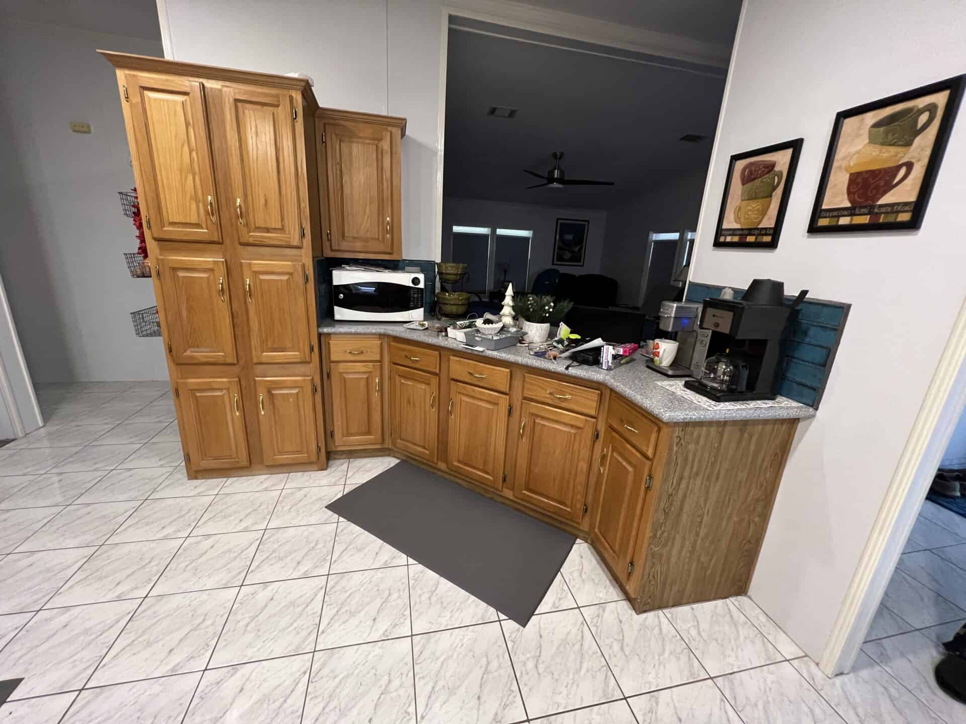 Wood-paneled kitchen corner with upper and lower cabinets, a microwave, coffee maker, and assorted items on the counter; white tiled floor.