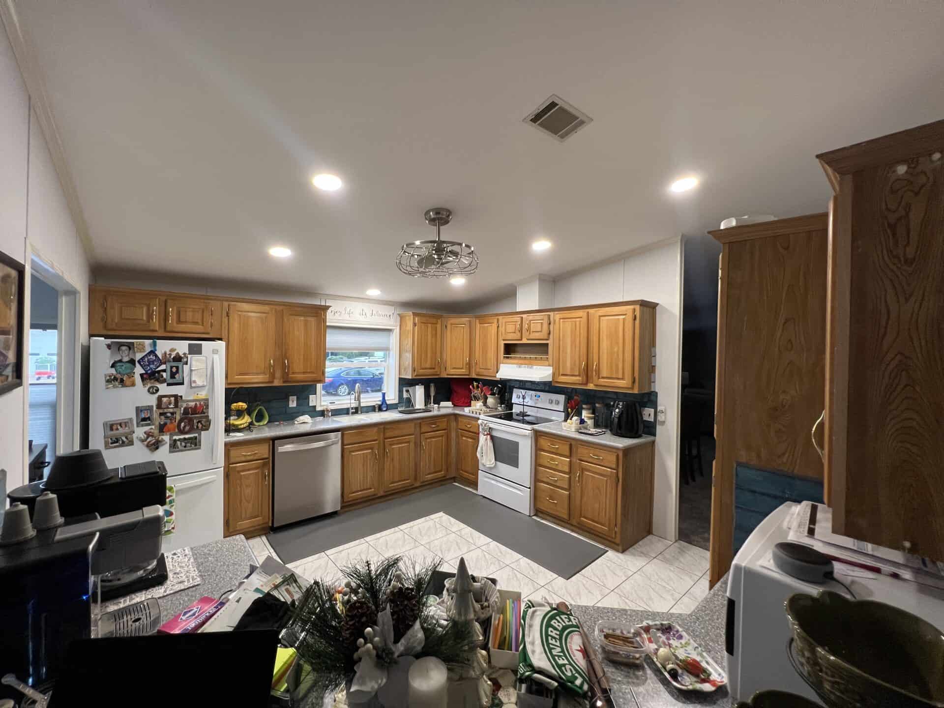 A cozy kitchen with wooden cabinets and a white tile floor. The fridge is adorned with family photos. A window above the sink lets in light, creating a warm, inviting atmosphere.