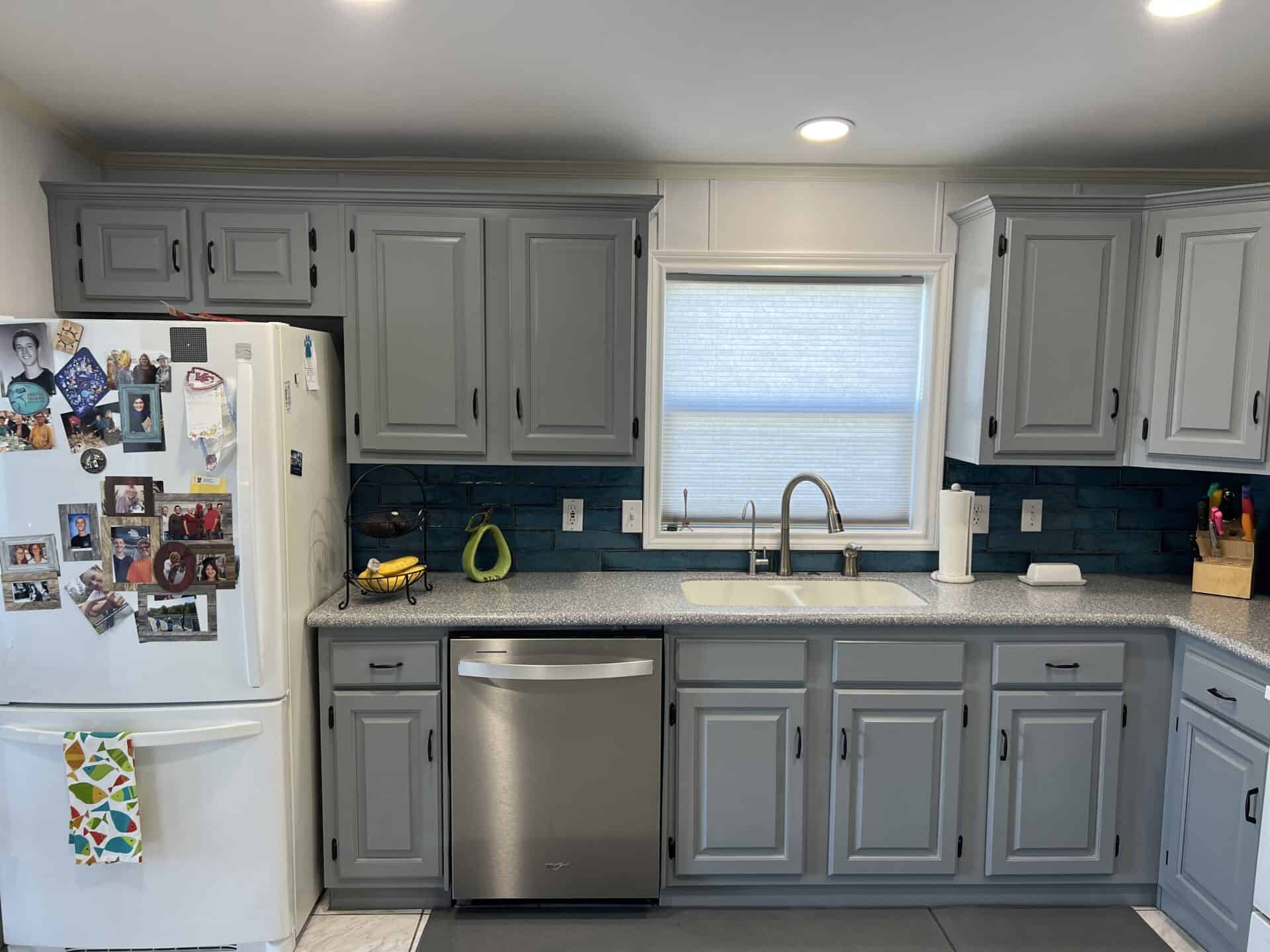 Grey kitchen with cabinets and a window above the sink. A white fridge displays various photos, while colorful utensils are on the counter. Bright, cozy atmosphere.