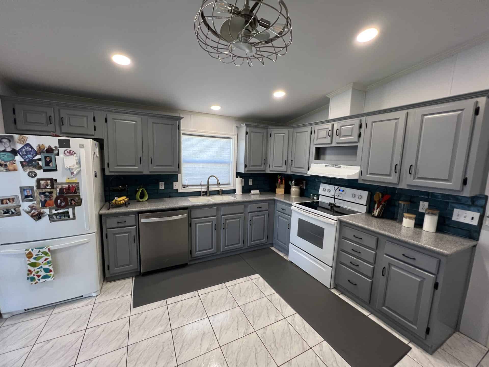 Modern kitchen with gray cabinets, white appliances, and tiled floor. Photos adorn the fridge. Bright lighting creates a clean, cozy atmosphere.