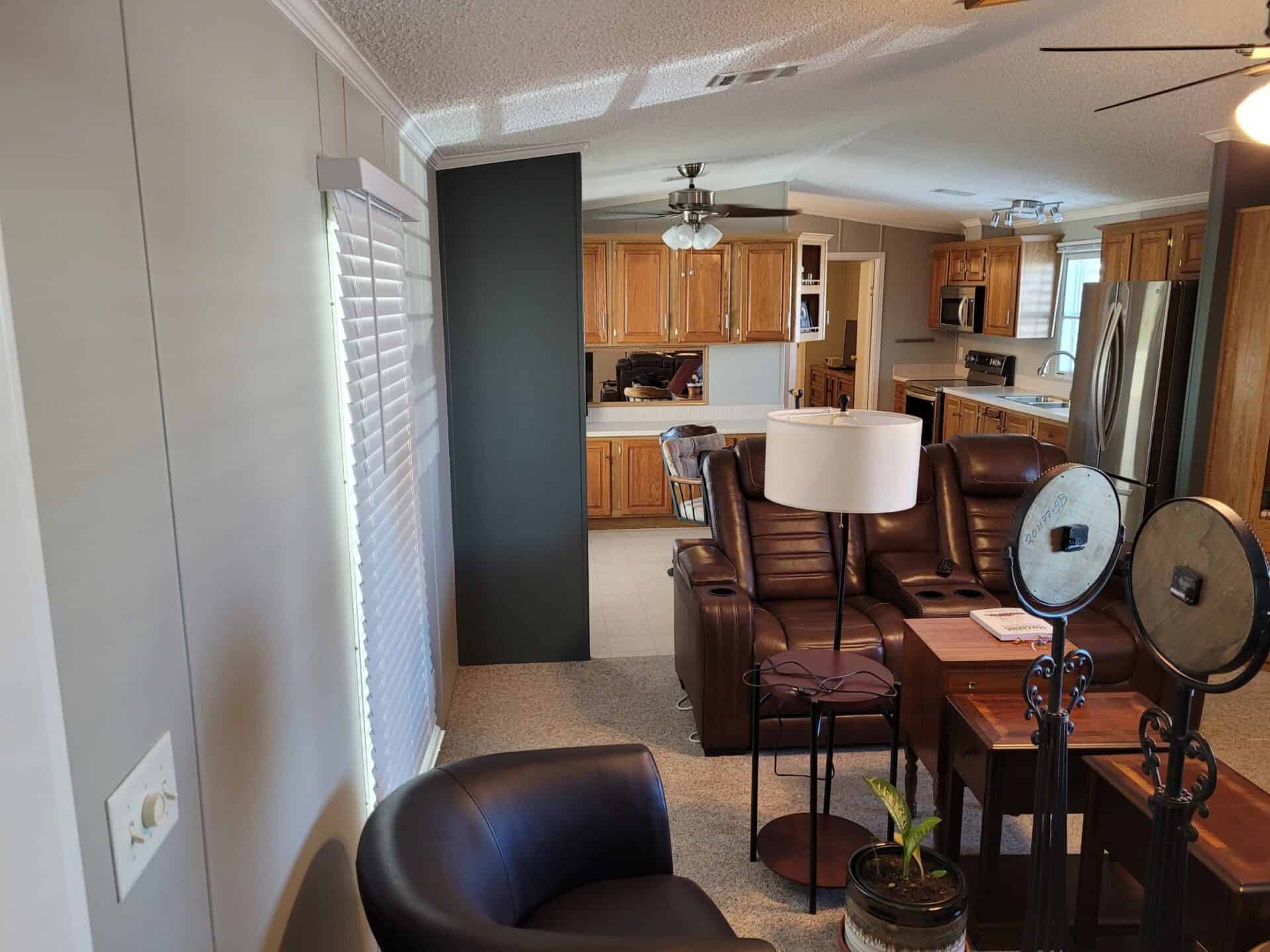 Modern living room with brown leather couches and a round black chair. A floor lamp and potted plant are nearby. Kitchen with wooden cabinets in the background. Cozy and inviting atmosphere.