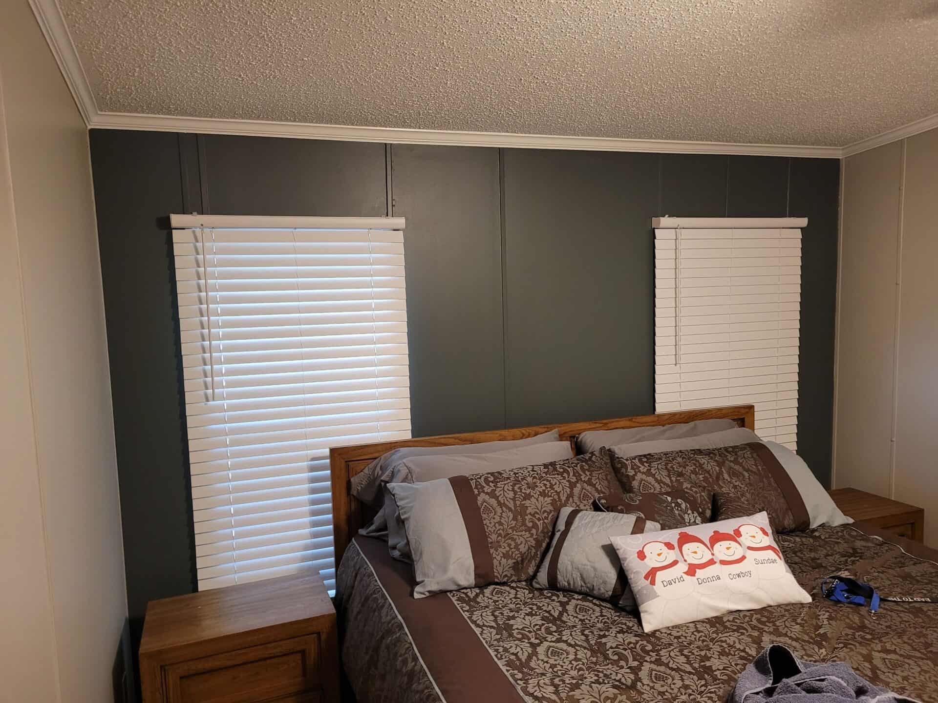 Cozy bedroom with earthy tones features a wooden bed against a gray accent wall. Two windows with white blinds flank the bed, and a playful pillow adds charm.