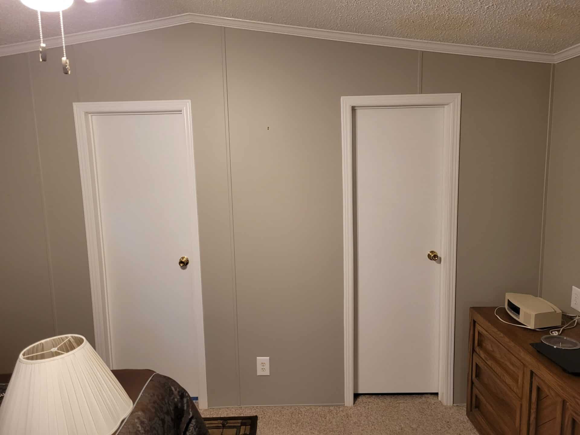 Room interior with two closed white doors on a gray wall. A wooden dresser with an electronic device is to the right. A white lamp is in the foreground.