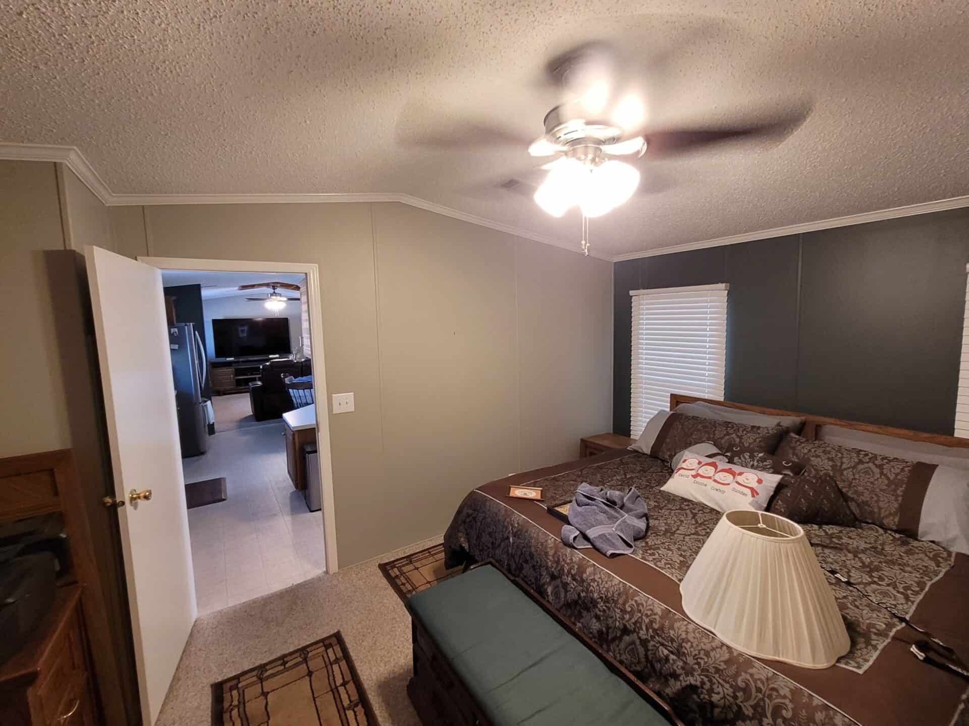 Cozy bedroom with a ceiling fan, large bed with patterned bedding, and decorative pillows. Open door reveals a view of a living room area.