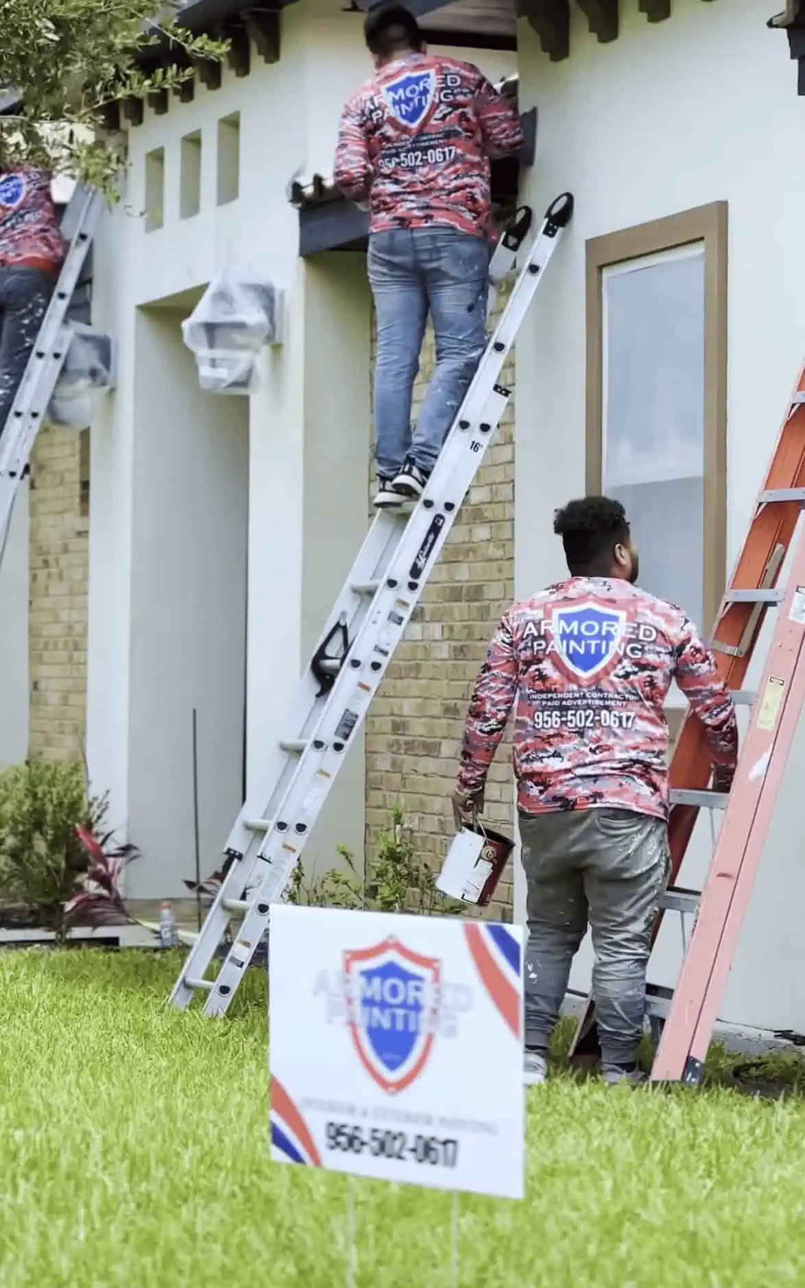 Two workers in red camo uniforms stand on ladders, painting a house exterior. A yard sign with "Armored Painting" is visible in the foreground.