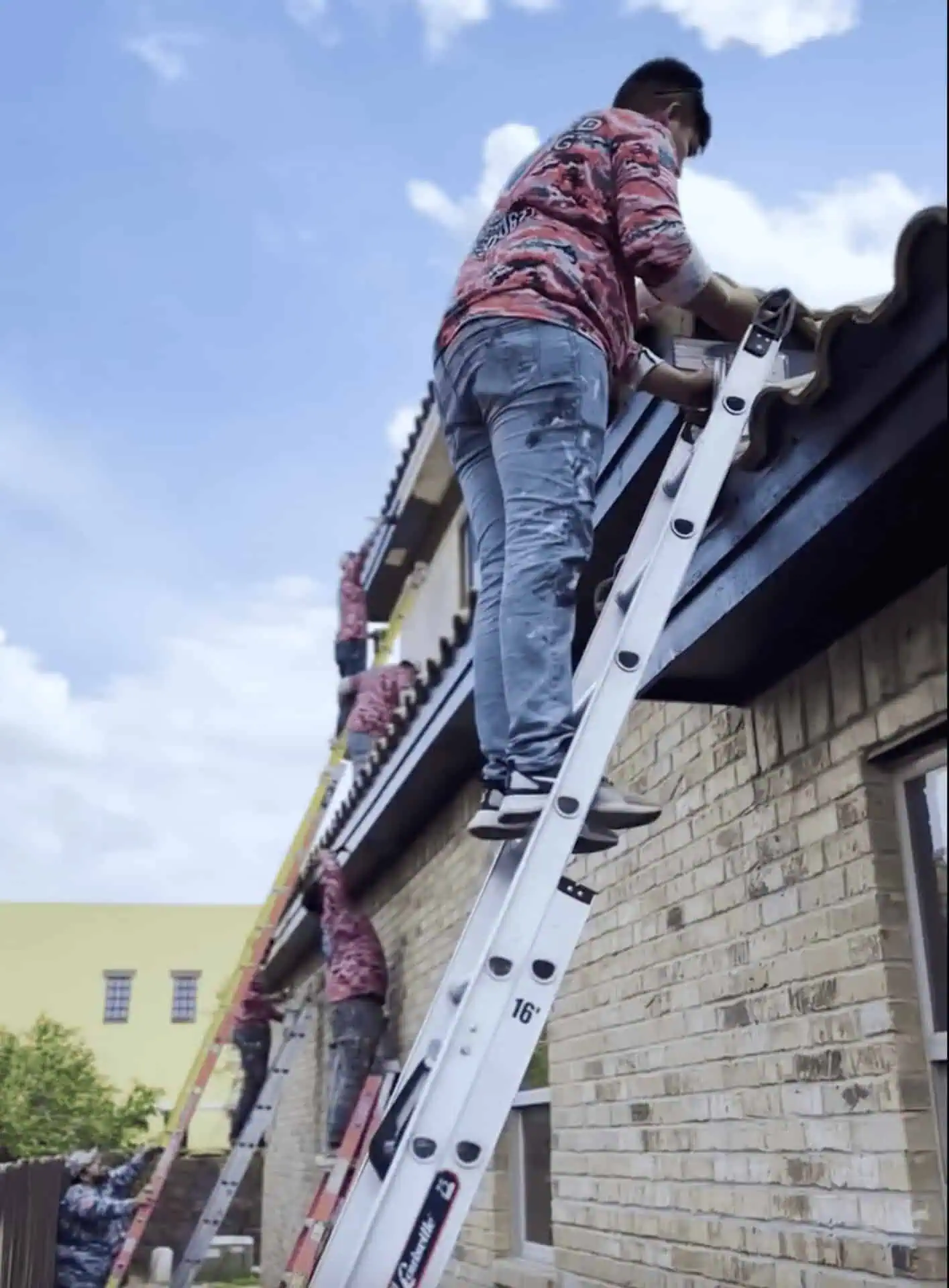 Three workers in red shirts stand on ladders, repairing a roof on a brick building. The sky is clear and blue, suggesting a calm work environment.