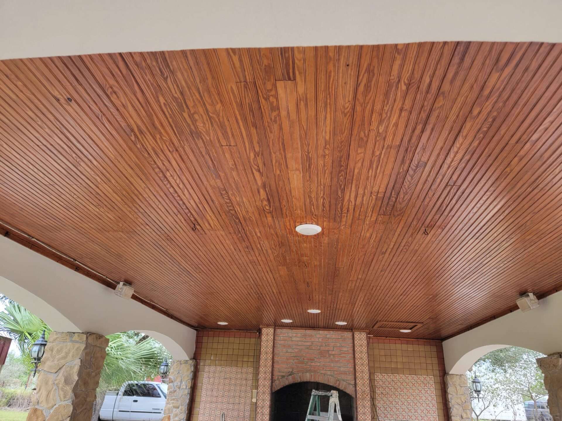 Polished wooden ceiling with embedded lights in a covered outdoor area. Stone pillars and arches lead outside, with a truck visible in the background.