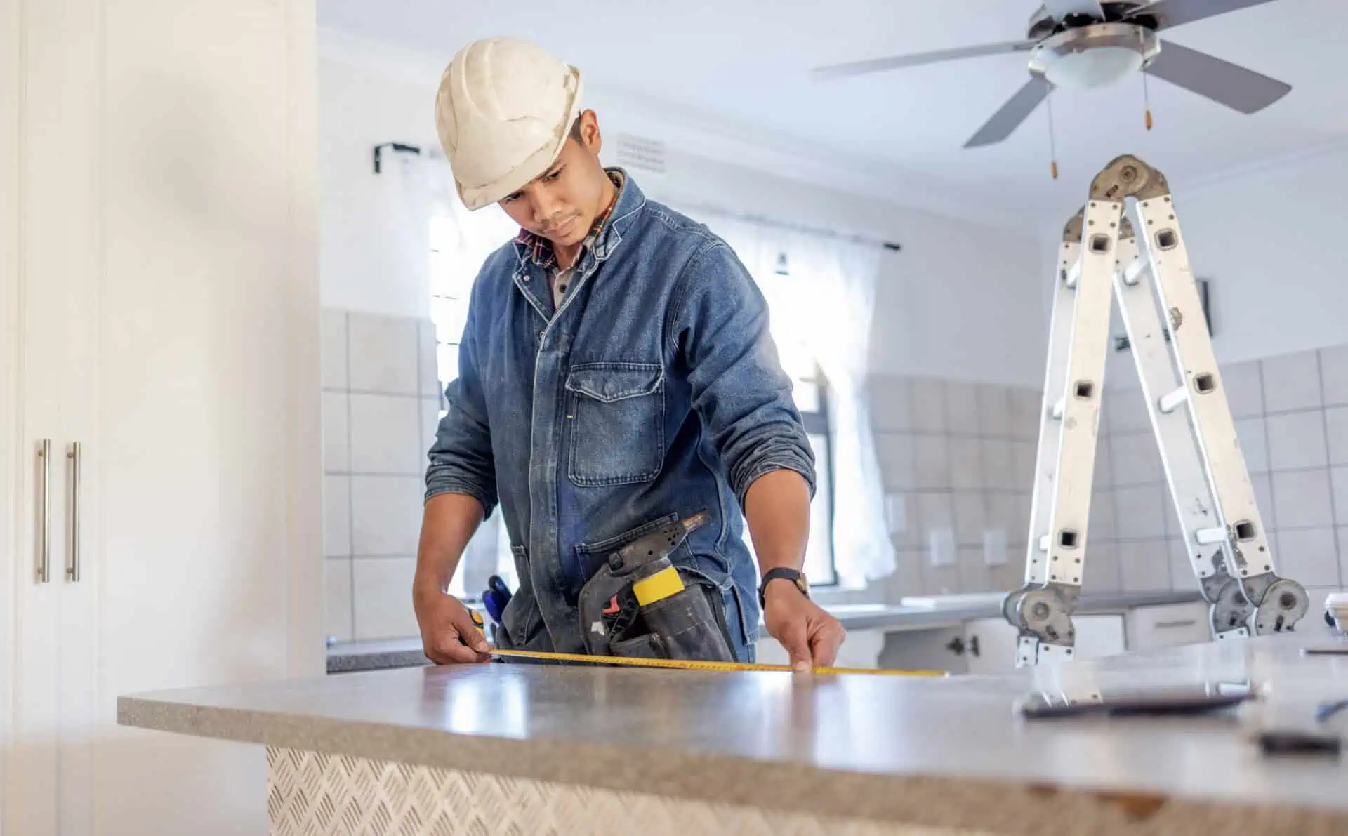 A construction worker in a denim shirt and hard hat measures a kitchen countertop with a tape measure, with focus and precision. A ladder and ceiling fan are visible in the background, creating a work-in-progress atmosphere.