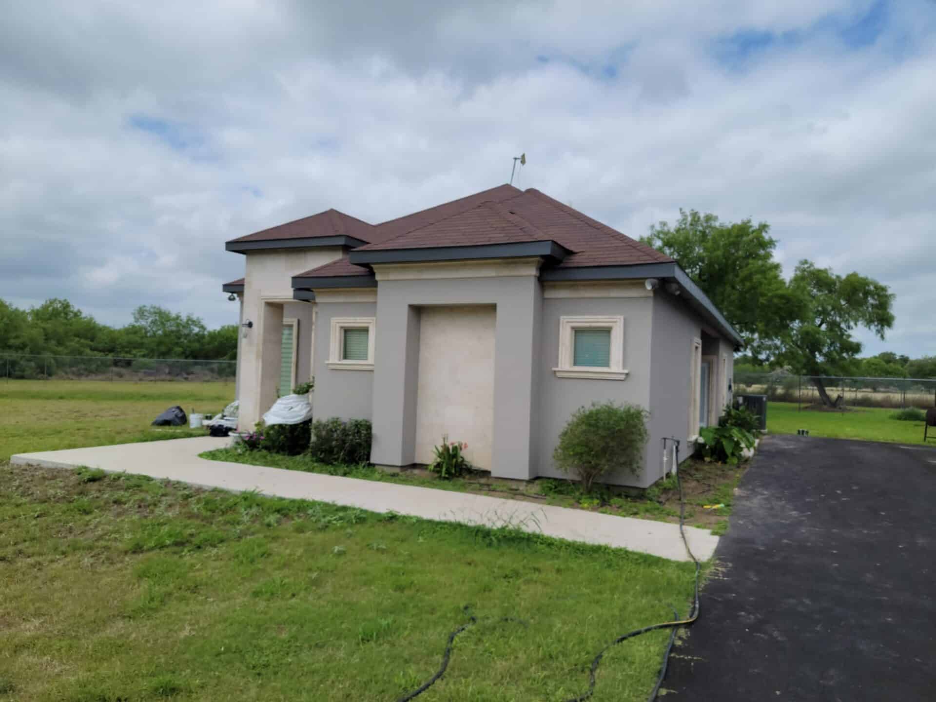 Small, beige house with a brown roof, surrounded by a lush green lawn and trees under a cloudy sky. Sidewalk and driveway lead to the entrance.