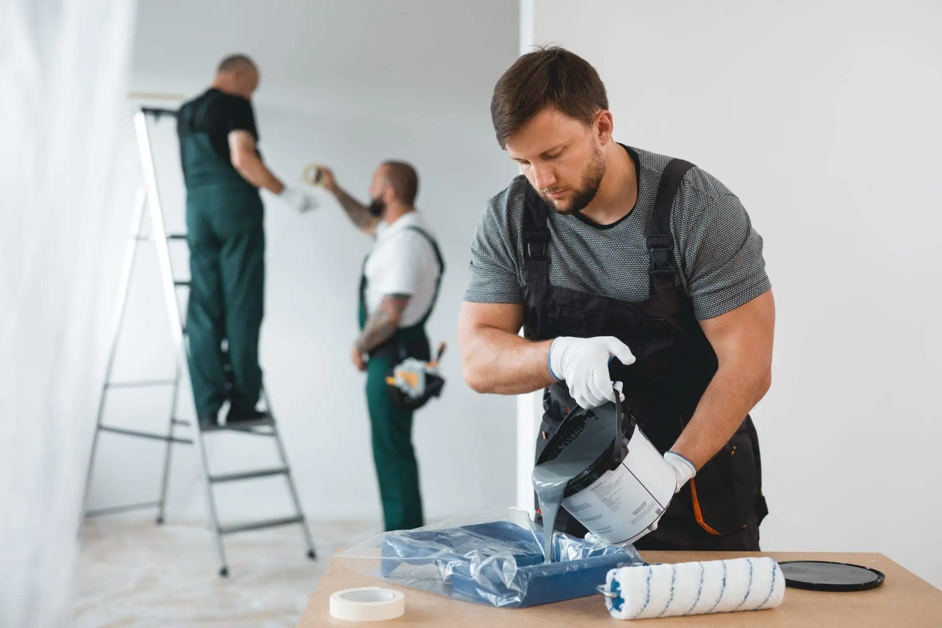A man in work overalls pours paint into a tray, focused and working carefully. Two others in similar attire work on a ladder, painting a room in soft tones.