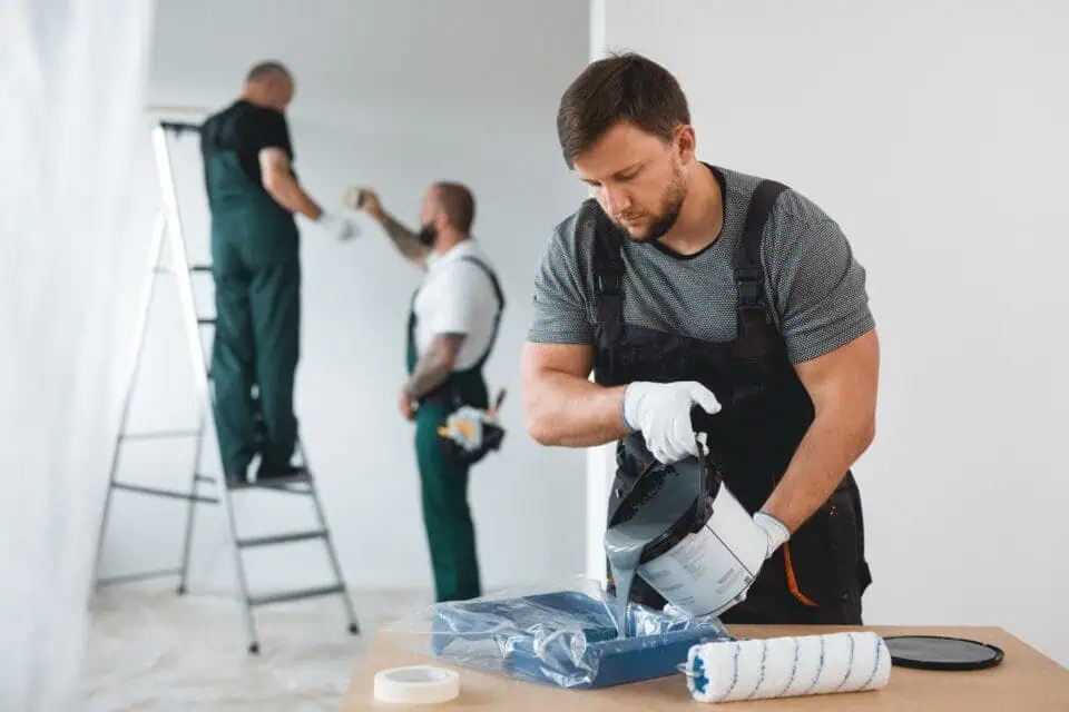 A man in work overalls pours paint into a tray, focused and working carefully. Two others in similar attire work on a ladder, painting a room in soft tones.