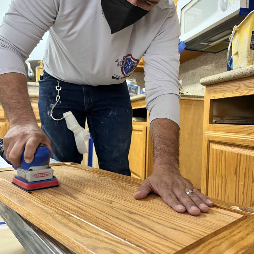A person wearing a red cap and black mask sands a wooden cabinet door in a kitchen. They focus intently, creating a sense of concentration.