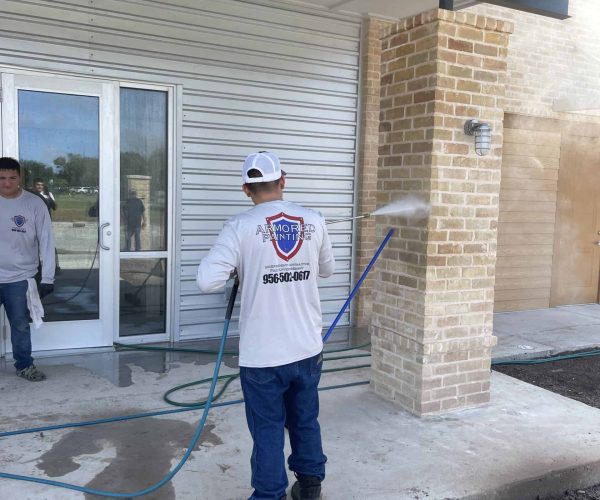 A person in a white shirt and cap is pressure washing a concrete walkway near a brick column. Another person stands nearby. The setting appears to be commercial.