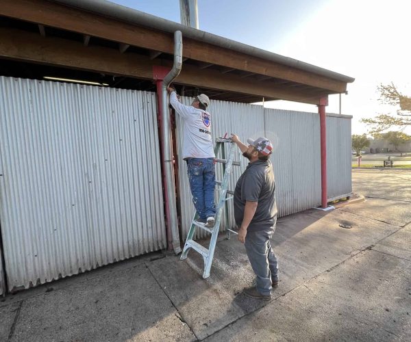 Two men work on a corrugated metal building. One stands on a ladder adjusting a pipe, while the other holds the ladder. The setting sun casts long shadows.