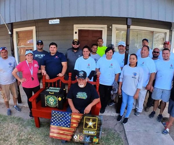 A group of 17 people stands in front of a house, smiling. An American flag waves from the porch. The mood is joyful and united, with military-themed items displayed.