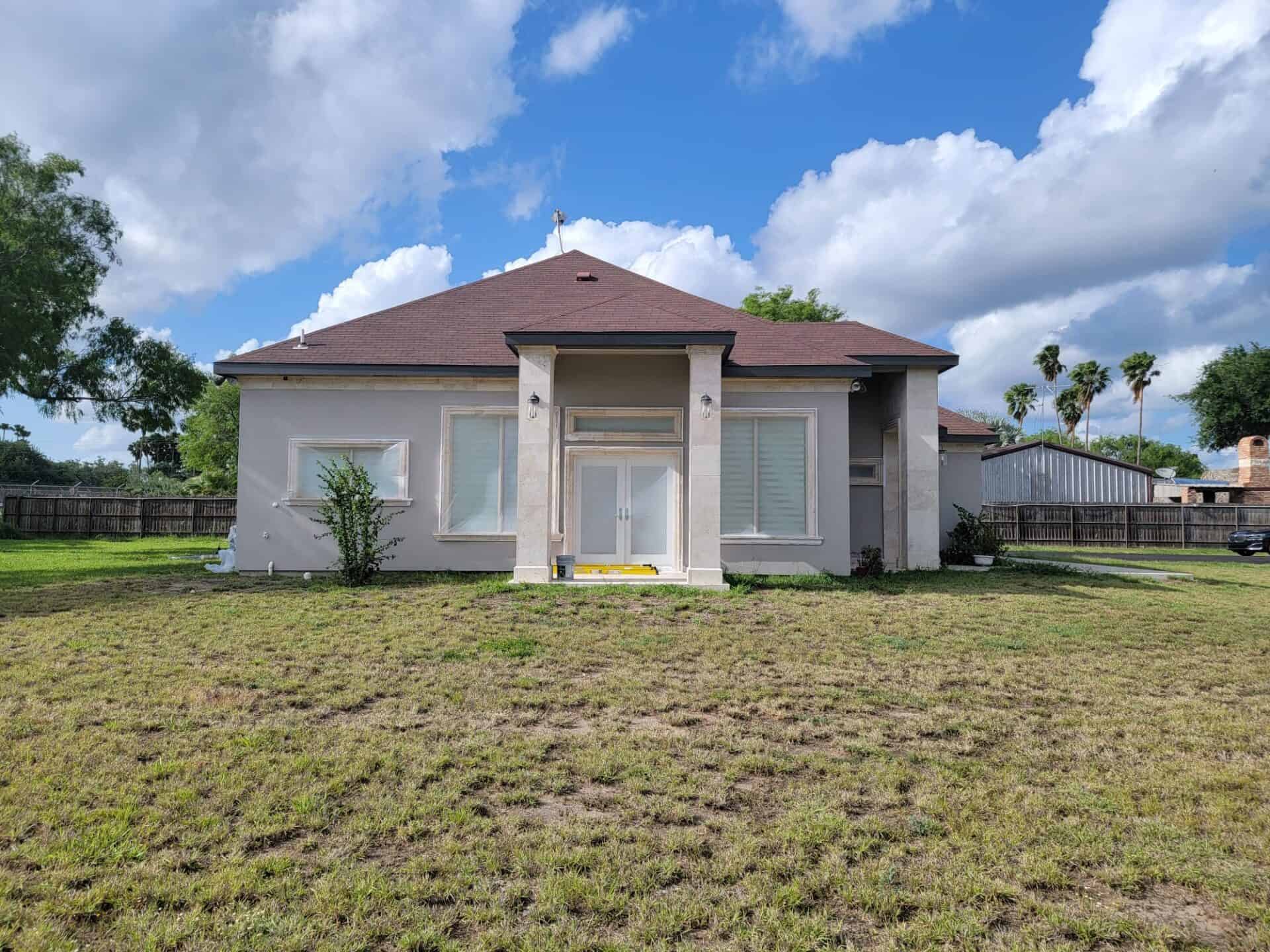Single-story house with a brown roof and light gray walls, set against a blue sky with clouds. The yard is grassy with a few plants. Quiet, suburban vibe.