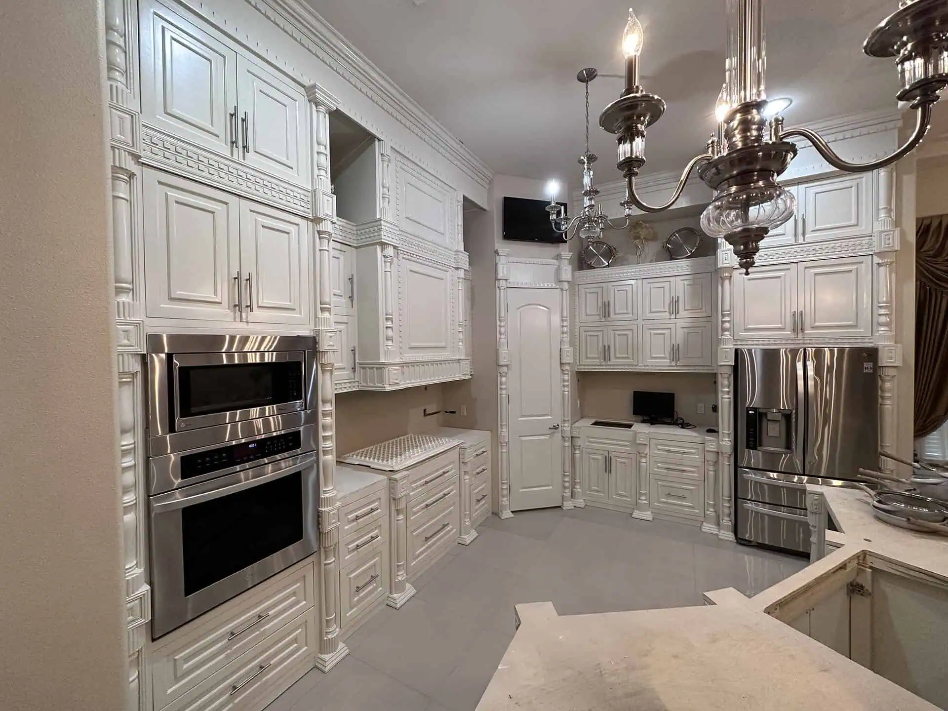 Elegant kitchen with ornate white cabinetry, a stainless-steel double oven, and fridge. A chandelier adds a luxurious touch to the spacious setting.