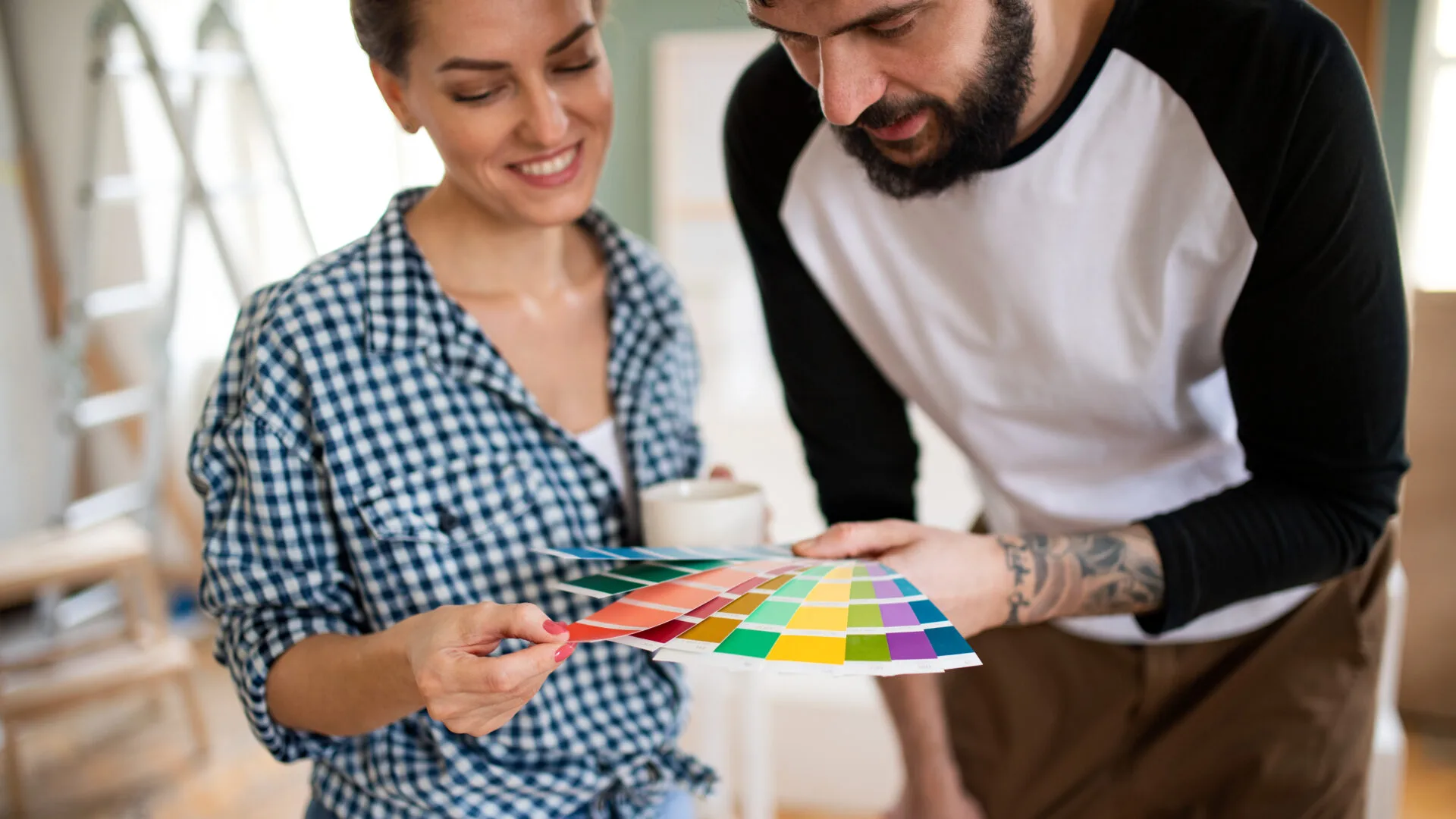 A smiling woman in a checkered shirt and a bearded man in a baseball tee examine colorful paint swatches together, conveying creativity and collaboration.