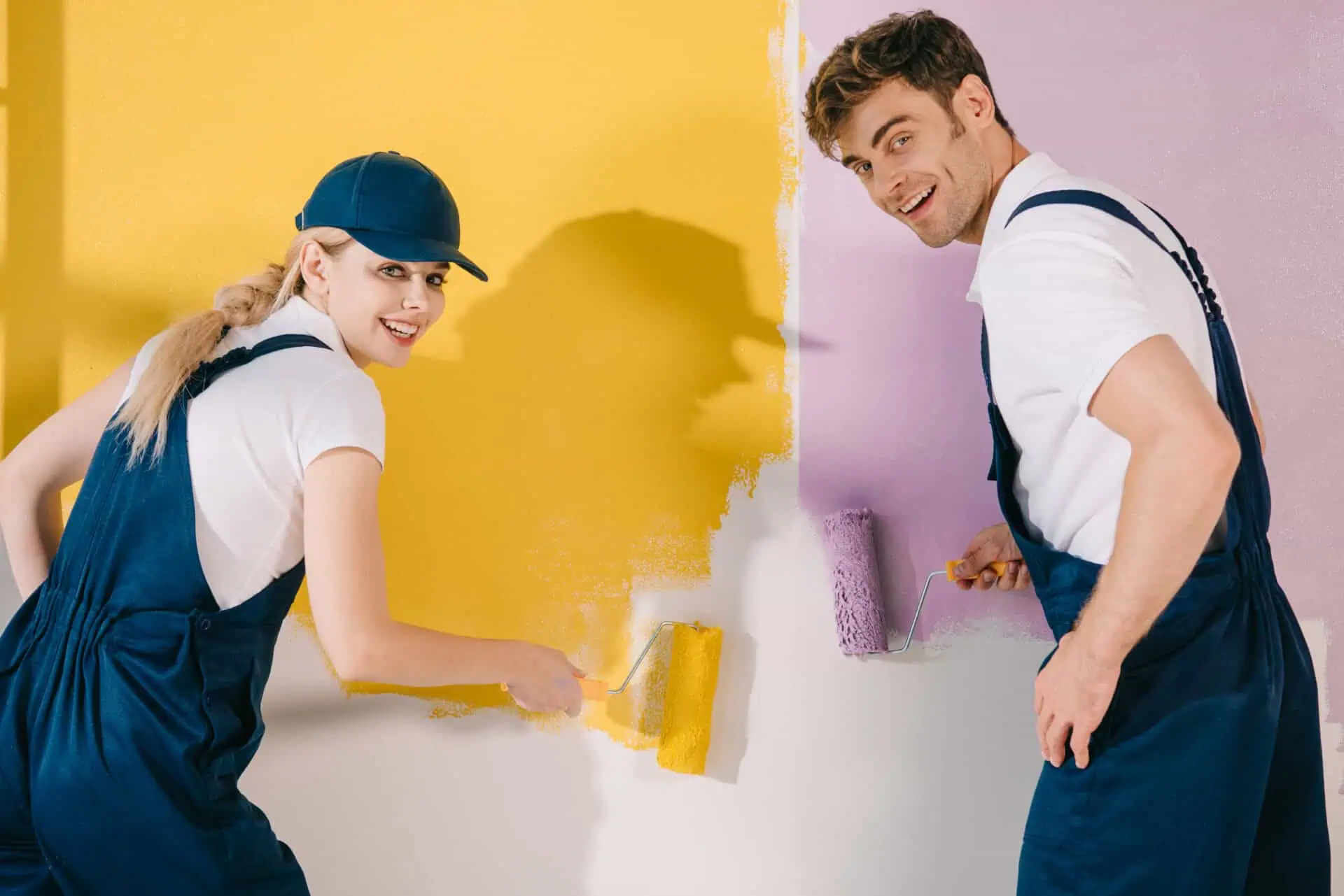 Two people in blue overalls cheerfully paint a wall, one with yellow and the other with purple, creating a vibrant and playful contrast.