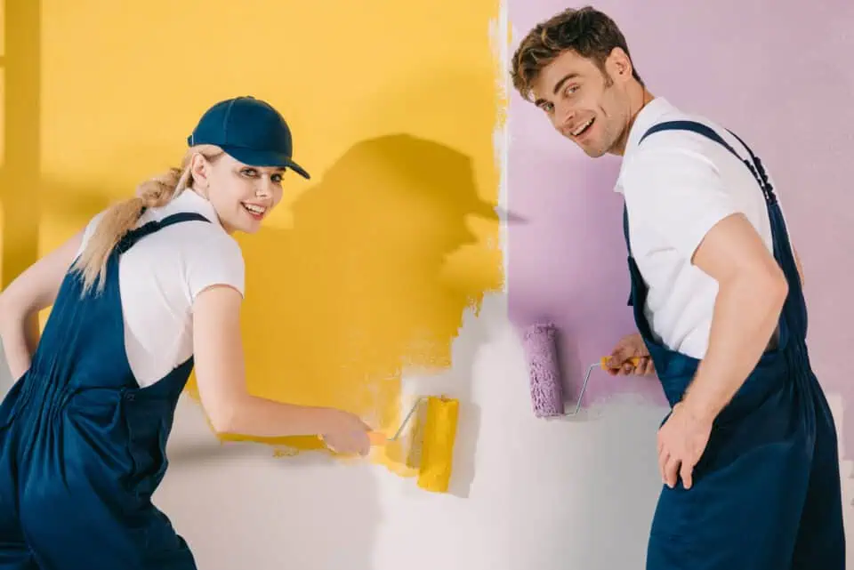 Two people in blue overalls cheerfully paint a wall, one with yellow and the other with purple, creating a vibrant and playful contrast.